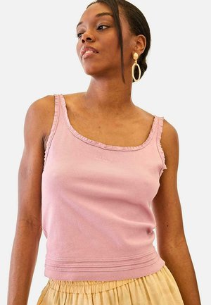 Mujer de cabello oscuro con grandes pendientes de aro dorados, una blusa sin mangas rosa y una falda amarillo claro, mirando hacia un lado sobre un fondo blanco.