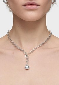 Collar de cadena de plata con eslabones ovalados, un colgante esférico central y una pequeña etiqueta. Textura lisa con un acabado pulido.