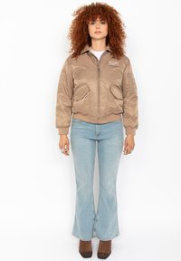 Braune Satin-Bomberjacke mit Reißverschlusstaschen und gerippten Bündchen, getragen über einem weißen Oberteil und hellblauen Schlagjeans, kombiniert mit braunen High Heels.