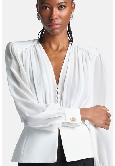 Femme aux cheveux tressés portant une blouse blanche à manches longues bouffantes, boutonnée devant, avec des boutons décoratifs aux poignets, posant bras croisés.