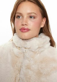 Manteau en fausse fourrure de couleur crème, avec un col haut, une texture douce et une ceinture avec des boutons dorés pour la fermeture.