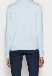 Persona vistiendo un suéter de manga larga azul claro con puños acanalados y jeans azul oscuro, mostrado de espaldas contra un fondo liso.