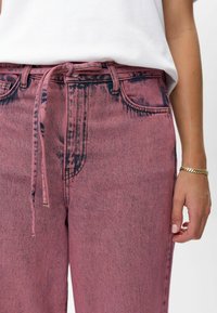 Ružové denimpantalóny s vysokým pásom, so stiahnuteľným opaskom, prednými vreckami a jemne vyblednutou textúrou. Tkanina vykazuje jemné farebné variácie.