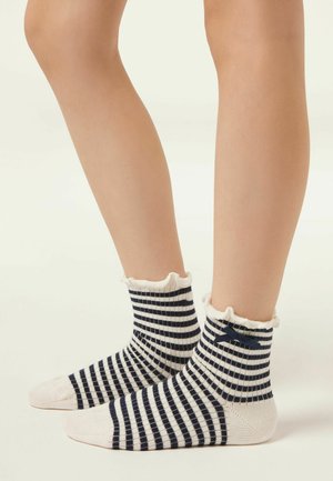 Beine, die marineblaue und weiße gestreifte Knöchelsocken mit Rüschensaum und kleinem marineblauem Schleifchen tragen, stehen auf einer hellen Oberfläche.