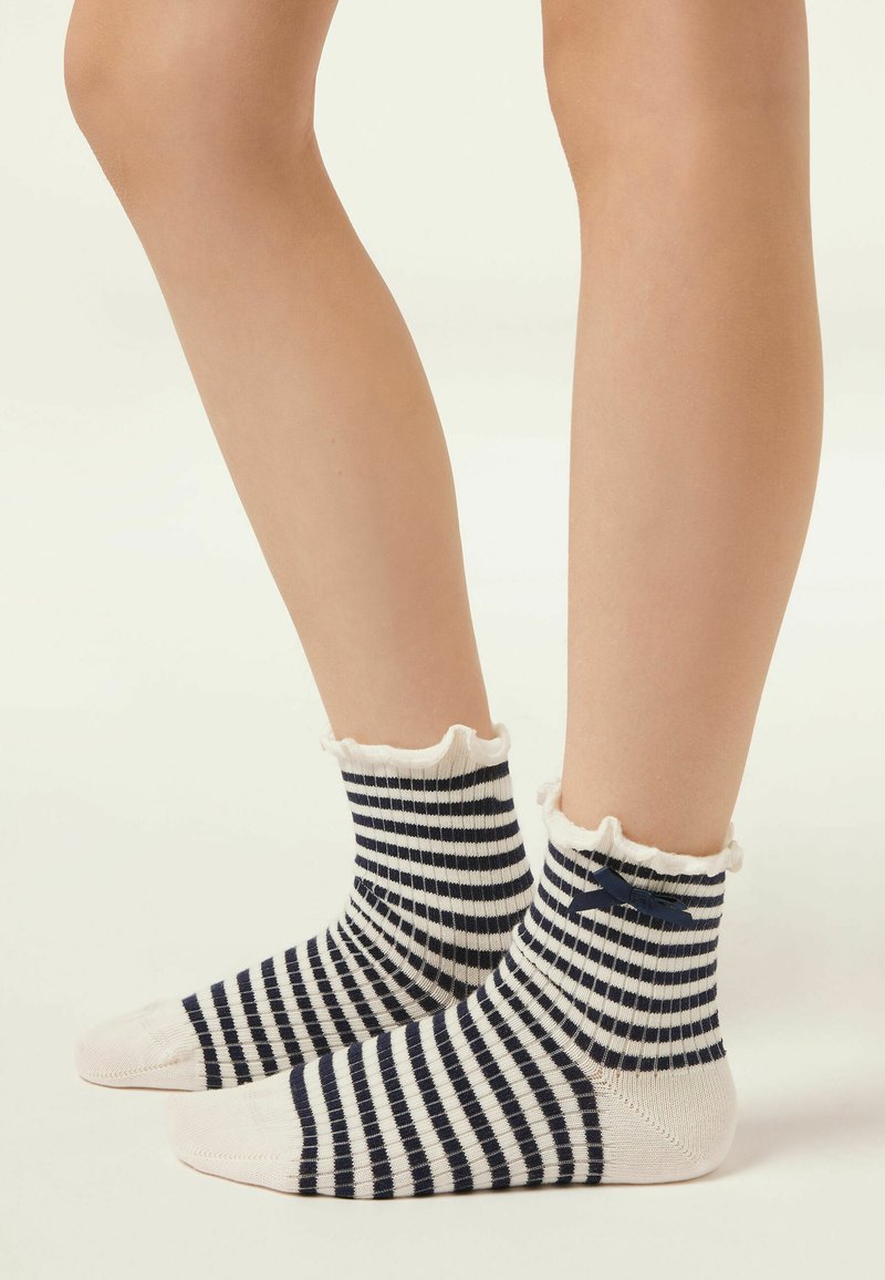 Jambes portant des chaussettes courtes rayées bleu marine et blanc avec des bords à volants et un petit nœud bleu marine, debout sur une surface de couleur claire.