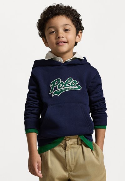 Sweatshirt marine avec des accents verts, arborant un logo "Polo Ralph Lauren". Superposé à une chemise blanche à col et associé à un pantalon kaki.