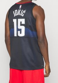 Czarny koszulkowy strój do koszykówki z białym napisem "JOKIĆ" i numerem "15". Posiada niebieskie akcenty i czerwony dekolt. Widoczna fakturowana tkanina.