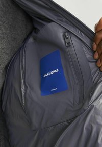 Grå indvendig foring af en jakke med et blåt JACK & JONES-label, med en lynlåslomme og glat tekstur.
