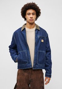 Jachetă din denim cu guler din catifea maro, fermoar în față și două buzunare mari. Materialul albastru are o textură estompată. Croi lejer, fără căptușeală.