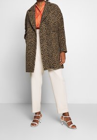 Cappotto a stampa leopardata, design oversize, colori beige e nero, tessuto strutturato, abbinato a pantaloni crema e tacchi bianchi con cinturini.