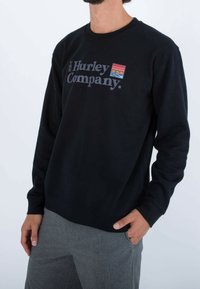 Černá mikina s dlouhými rukávy, žebrovanými manžetami a kulatým výstřihem. Na přední straně má vícero barevné grafické logo.