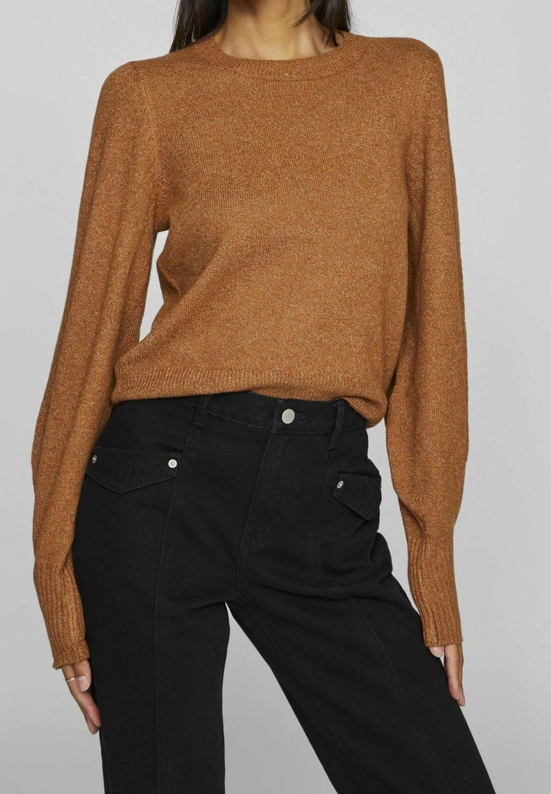 Femme portant un pull en maille marron à manches longues, rentré dans un pantalon noir taille haute avec des poches à rabat à l'avant.