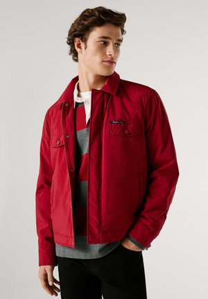 Ung mand iført en rød jakke over en grå og rødstribet skjorte, med den ene hånd i lommen, kiggende let til siden.