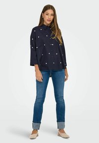 Navy button-up shirt with white heart-shaped patterns, three-quarter sleeves, and a relaxed fit, paired with blue cuffed jeans and beige flats.