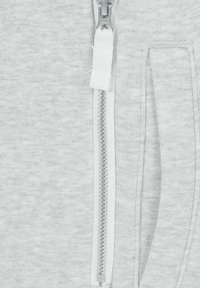 Sudadera con cremallera de color gris claro, que presenta una textura de tela suave, una cremallera blanca y un bolsillo lateral. Los dientes de la cremallera son claramente visibles.