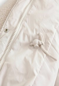 Veste zippée blanche avec une texture en sherpa peluche, un tissu extérieur lisse, un cordon de serrage ajustable avec un embout en plastique et un détail de pression en métal.