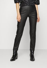 Musta tekonahka housut kapealla leikkauksella, joissa on siistit linjat, huomaamaton ompelu, ja jotka on yhdistetty mustiin korkokenkiin.