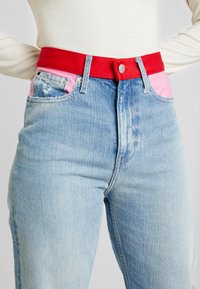 Höga ljusblå jeans med rosa fickfoder och ett klart rött bälte, bärs med en krämfärgad långärmad topp.