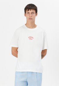 Witte katoenen T-shirt met korte mouwen, een ronde hals en een rode grafische tekstontwerp op de voorkant, casual gecombineerd met lichtblauwe broeken.