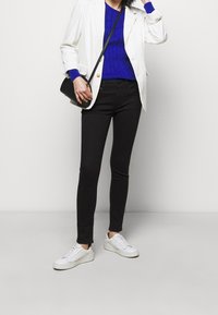 Vit kavaj över en blå kabelstickad tröja, kombinerad med svarta skinny jeans och vita sneakers. Inkluderar en svart axelremsväska.