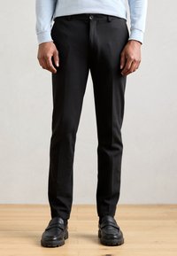 Pantalon noir avec une coupe ajustée, fabriqué à partir d'un tissu lisse. Présente une fermeture à bouton et des passants de ceinture, associé à des chaussures noires.