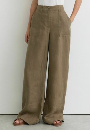 Vrouw draagt brede bruine broek met steekzakken aan de voorkant en knoopsluiting, gecombineerd met bruine sandalen en een wit topje.