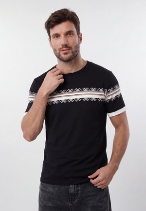 Muškarac s bradom u crnoj majici s bijelim i bež geometrijskim prugama preko prsa i rukava, podešava ovratnik, neutralna pozadina.