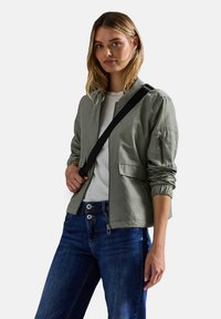 Donna che indossa una giacca bomber verde chiaro, una camicia bianca, jeans blu e una tracolla nera, in piedi contro uno sfondo bianco.