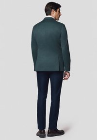 Ciemnozielona, dopasowana marynarka z pojedynczym rozcięciem, zestawiona z granatowymi spodniami. Dopełnieniem stroju są brązowe eleganckie buty. Gładka faktura tkaniny.