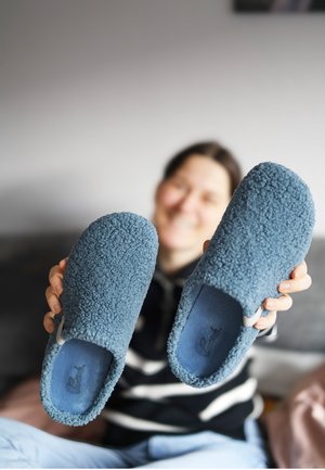 Persoon die een paar blauwe, pluizige sloffen binnenshuis vasthoudt, met de sloffen scherp in beeld en het gezicht vaag op de achtergrond.