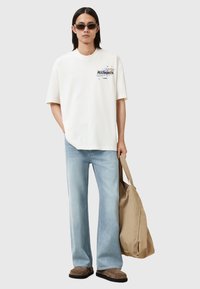 Camiseta de algodón blanca con estampado de logo en la parte delantera, jeans de mezclilla azul claro, zapatos marrones sin cordones y una tote bag beige. Textura suave, corte relajado.