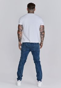 T-shirt bianco in cotone, jeans blu e sneaker bianche. Il T-shirt ha maniche corte, i jeans hanno tasche e una texture in denim sbiadito.