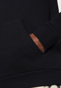 Svart sweatshirt med känguruficka och ribbade ärmslut. Har en brun läderdetalj på insidan av ärmsluten. Slät textur.