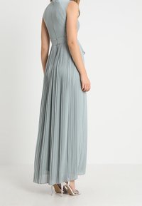 Maximekter i blødt, lys grå plisseret stof med en tætsiddende overdel, høj halsudskæring og en udvidet nederdel, parret med åbne hæle.