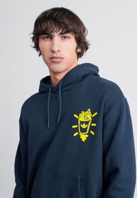Hanorac pulover de culoare albastru marin, cu buzunar frontal, având un grafic galben al unui recipient pentru cartofi prăjiți cu raze, realizat dintr-un material moale și texturat.
