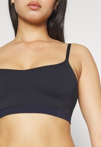 Svart bralette med justerbare, tynne stropper, en glatt tekstur og en bred kant nederst. Minimalistisk design, ingen synlige lukkinger.