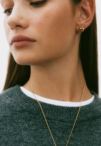 Kulla sõlmega kõrvarõngad modellil, kes kannab halli kampsunit ja valget sisepaela. Kaeluse ja kõrva detailide lähivõte.