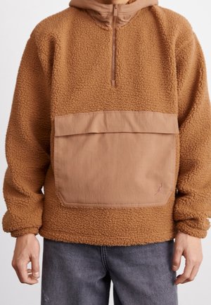 Pull-over en polaire marron avec une fermeture éclair devant, une grande poche avant à rabat, et une coupe décontractée portée avec un jean bleu.