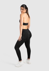 Ensemble de vêtements de sport noir comprenant un haut à col halter avec un dos ouvert et un legging taille haute, fabriqués en tissu lisse et extensible. Des baskets blanches viennent rehausser le look.