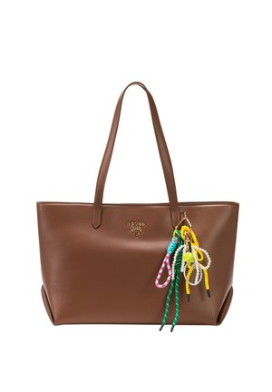 Bruine leren tote tas met twee handvatten en kleurrijke gevlochten sleutelhangers met kralen en een tennisbalbedeltje eraan bevestigd.