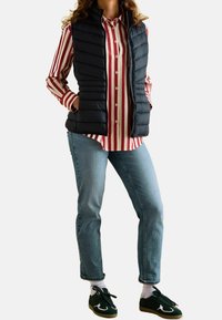 Czarna kamizelka puchowa nałożona na czerwono-białą koszulę w paski, w połączeniu z jasnoniebieskimi dżinsami i zielonymi trampkami z białymi akcentami.