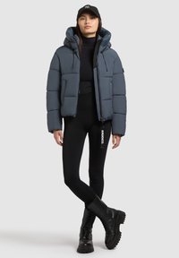 Pufferjack in donkergrijs met een capuchon, een ritssluiting aan de voorkant en elastische manchetten. Gekoeld met zwarte legging en zwart veterschoenen.