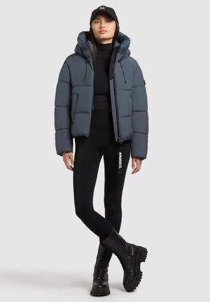 Puhasta jakna v temno sivi barvi s kapuco, z zadrgo spredaj in elastičnimi manšetami. V kombinaciji z črnimi legi in črnimi čevlji z vezalkami.