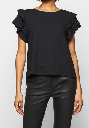 Osoba w czarnej bluzce z falbankami na rękawach i czarnych skórzanych spodniach, pokazana od ramion do bioder na jednolitym tle.