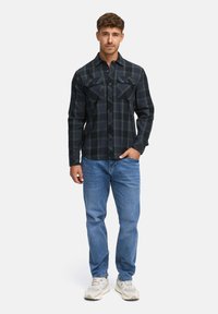 Schwarz-grünes karierter Flanellhemd mit Knopfdesign und zwei Brusttaschen, kombiniert mit hellblauen Jeans und beigen Sneakers.