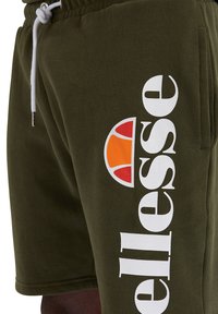Olivinės spalvos šortai pagaminti iš minkštos medžiagos, su baltu "ellesse" logotipu ir spalvinga puslankio akcentu raudonos ir oranžinės spalvų. Reguliuojama raištelio juosta.