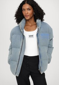 Gaišā zilā krāsā piepildīta džinsu jaka ar augstu apkakli, rāvējslēdzēja aizdari un zilo izšūto „Moschino Jeans” logo.