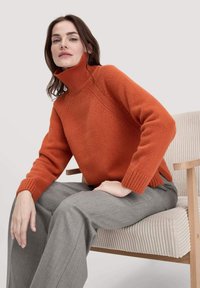 Helle orange Strickrollkragensweater mit gerippten Bündchen und Kragen, kombiniert mit hellgrauen Hosen. Die Textur wirkt weich und gestrickt.