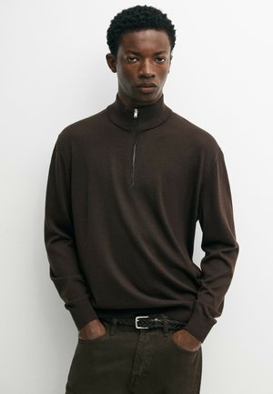 Ung mand stående med hænderne i lommerne iført en mørkebrun lynlås-sweater og mørke bukser mod en ensfarvet hvid baggrund.