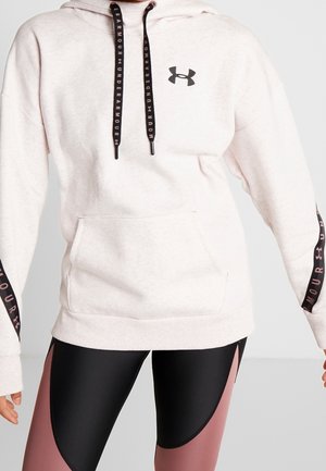 Vrouw draagt een lichtroze Under Armour hoodie met zwarte trekkoorden en zwarte en roze legging tegen een effen achtergrond.